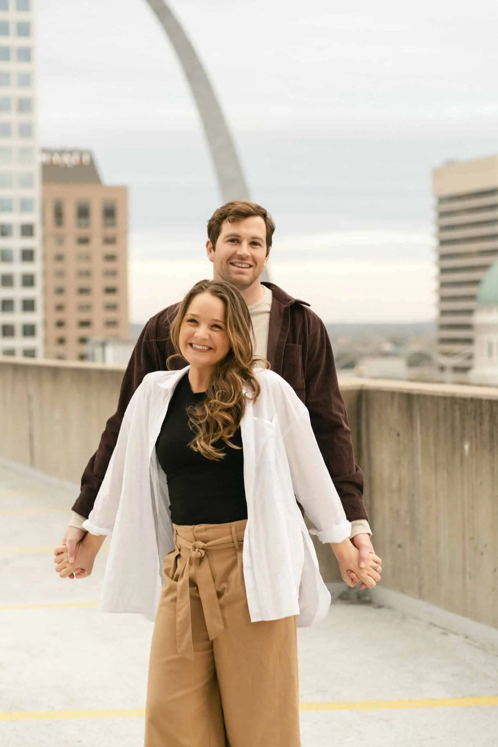 A couple smiling and holding hands on a rooftop in St. Louis with the Arch in the background, shot by Stacey Vandas Photography.