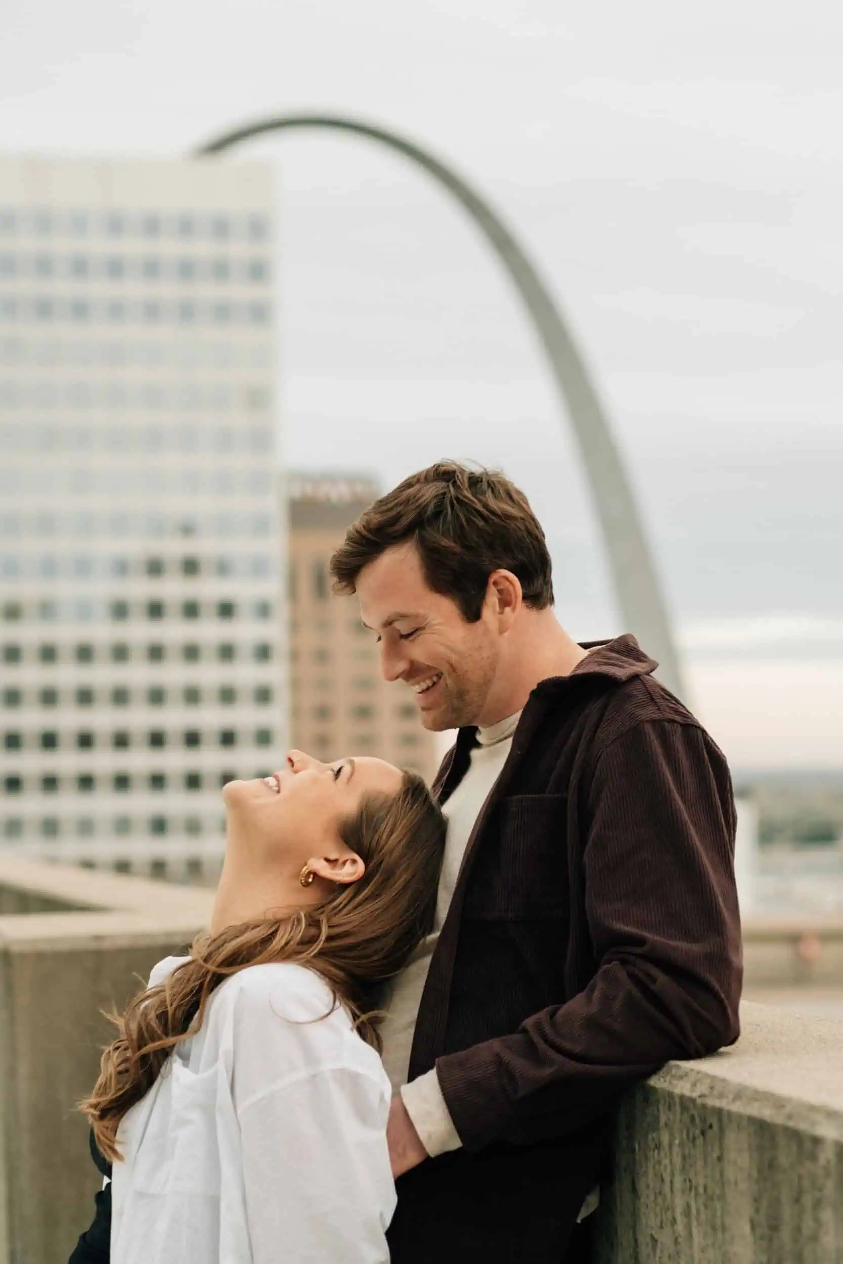 A couple leaning on the edge of a rooftop and smiling, with the Saint Louis Arch in the background, shot by Stacey Vandas Photography with a film style.
