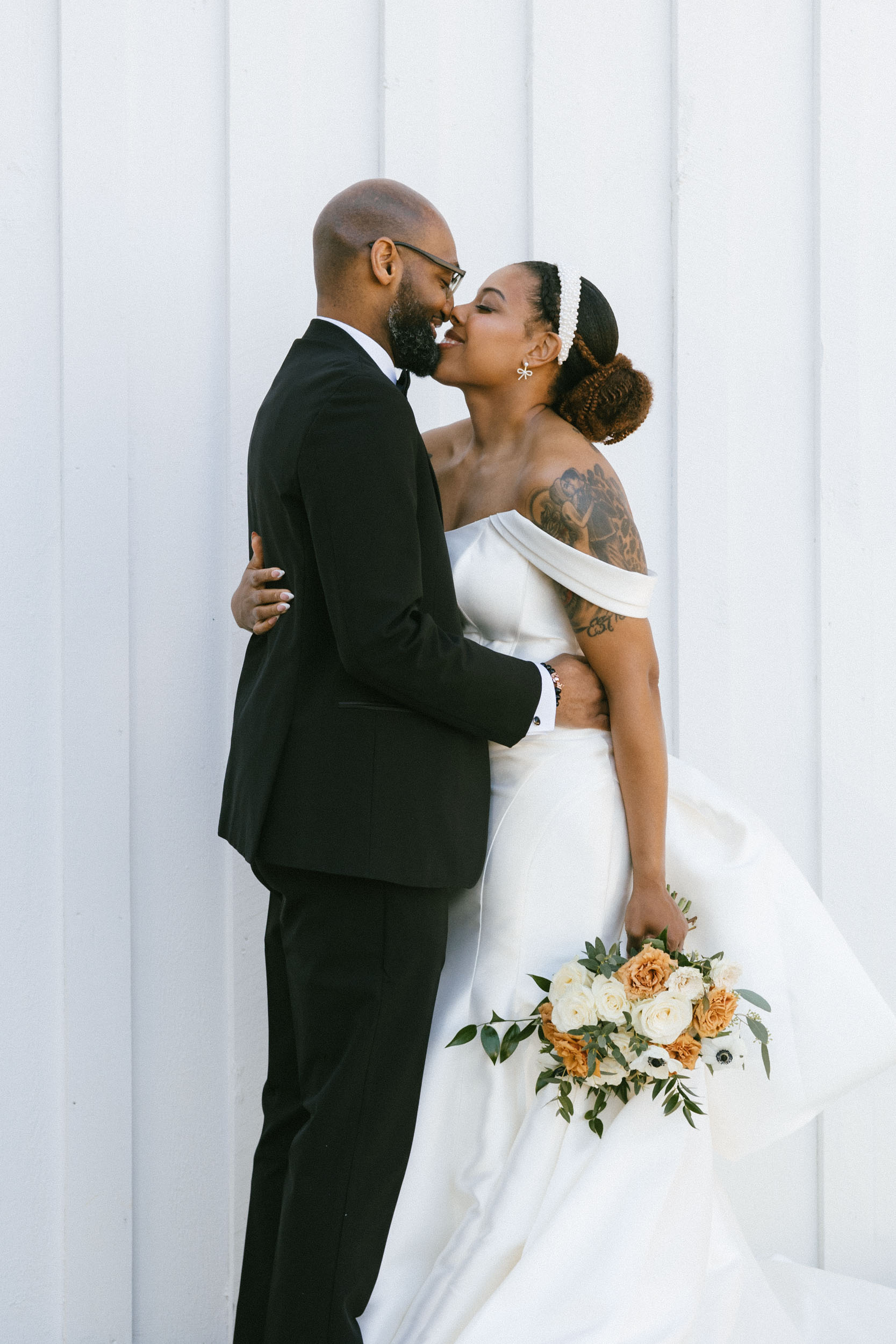 A couple embraces happily during an elopement wedding photoshoot in front of a white barn wall.