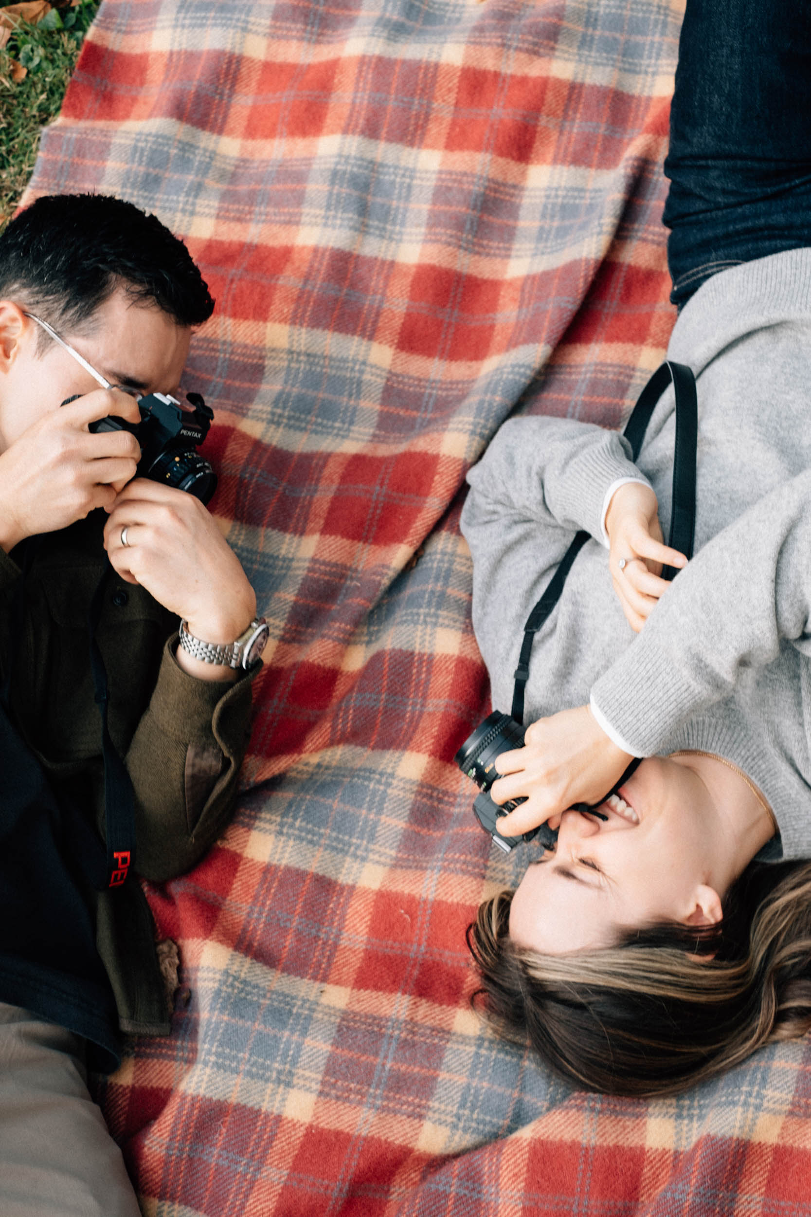 A couple lying on a plaid blanket and taking photos of one another with vintage film cameras, shot by Stacey Vandas Photography.