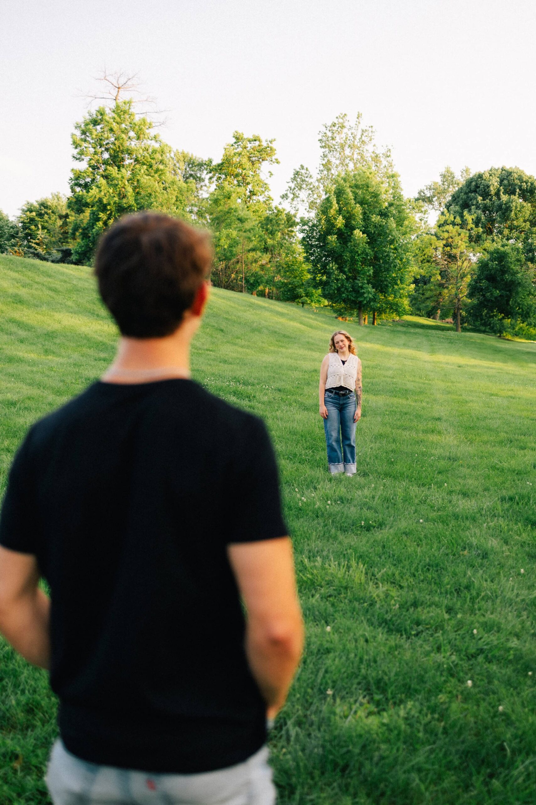 A man looks across a grassy field at his fiancée in an outdoor engagement photoshoot in Forest Park.