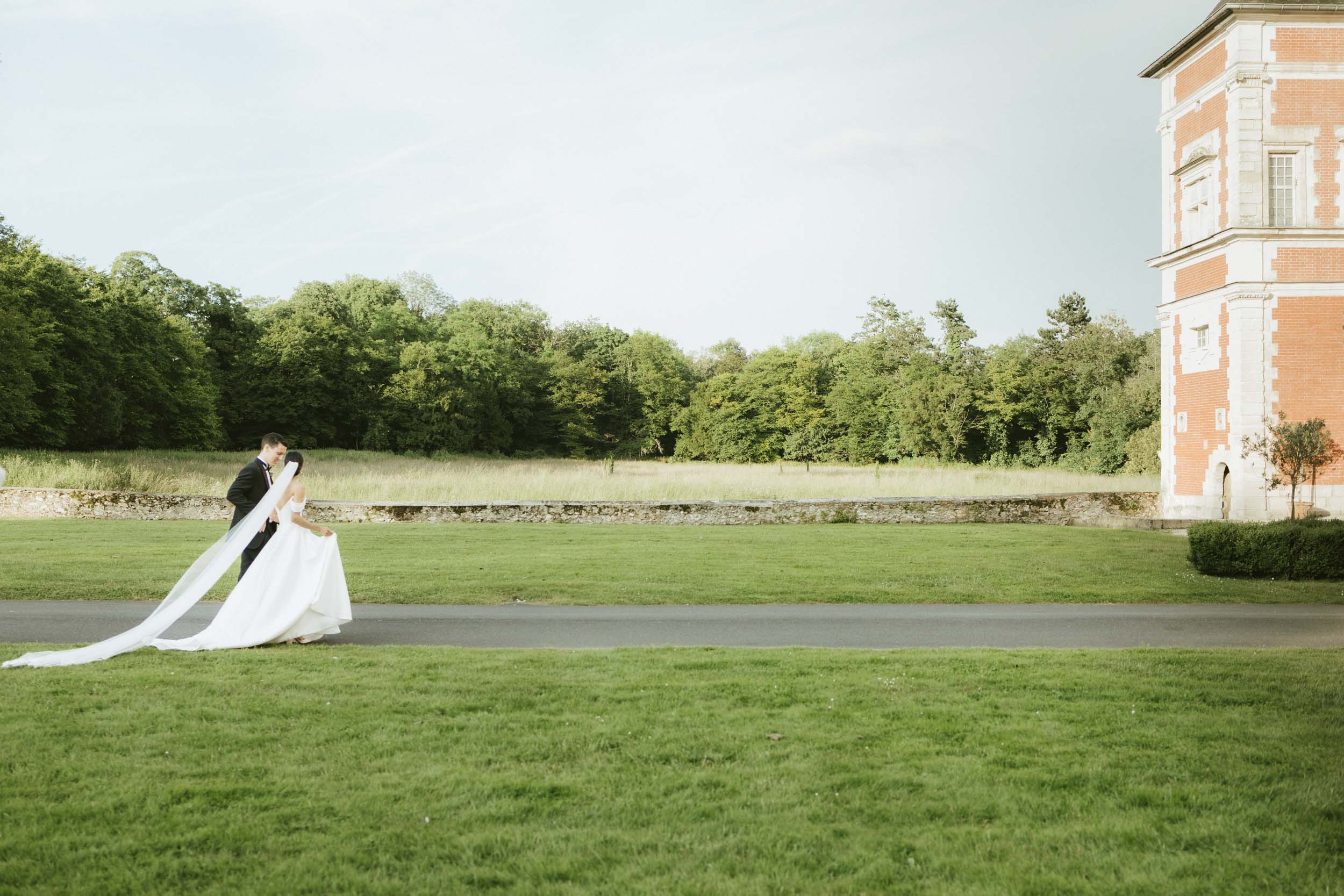 A bride and groom walk across a green lawn toward a château at their destination wedding in France.