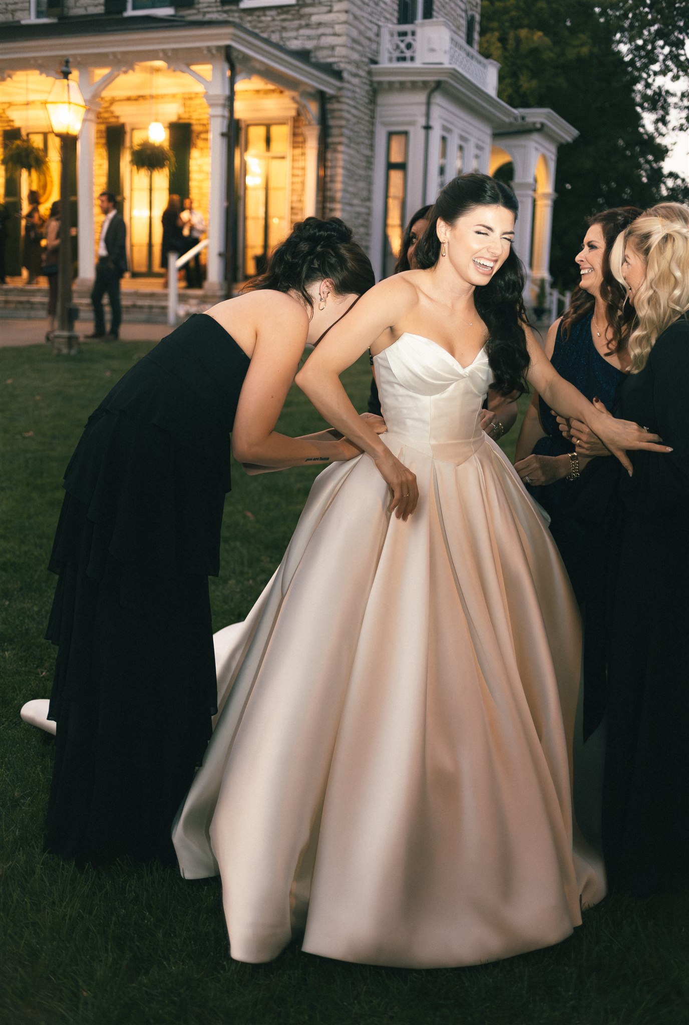 A bride laughs as her family bustles her dress outside her wedding venue in a documentary style wedding photo.