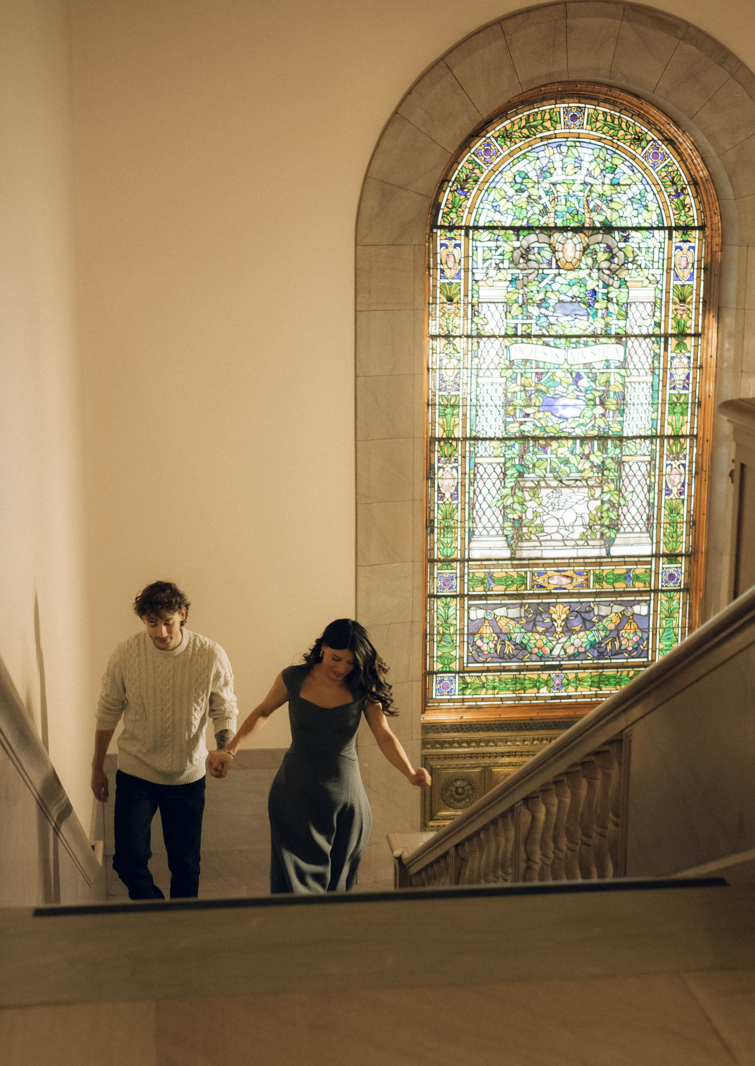 A couple runs up a staircase with a stained glass window in the background.