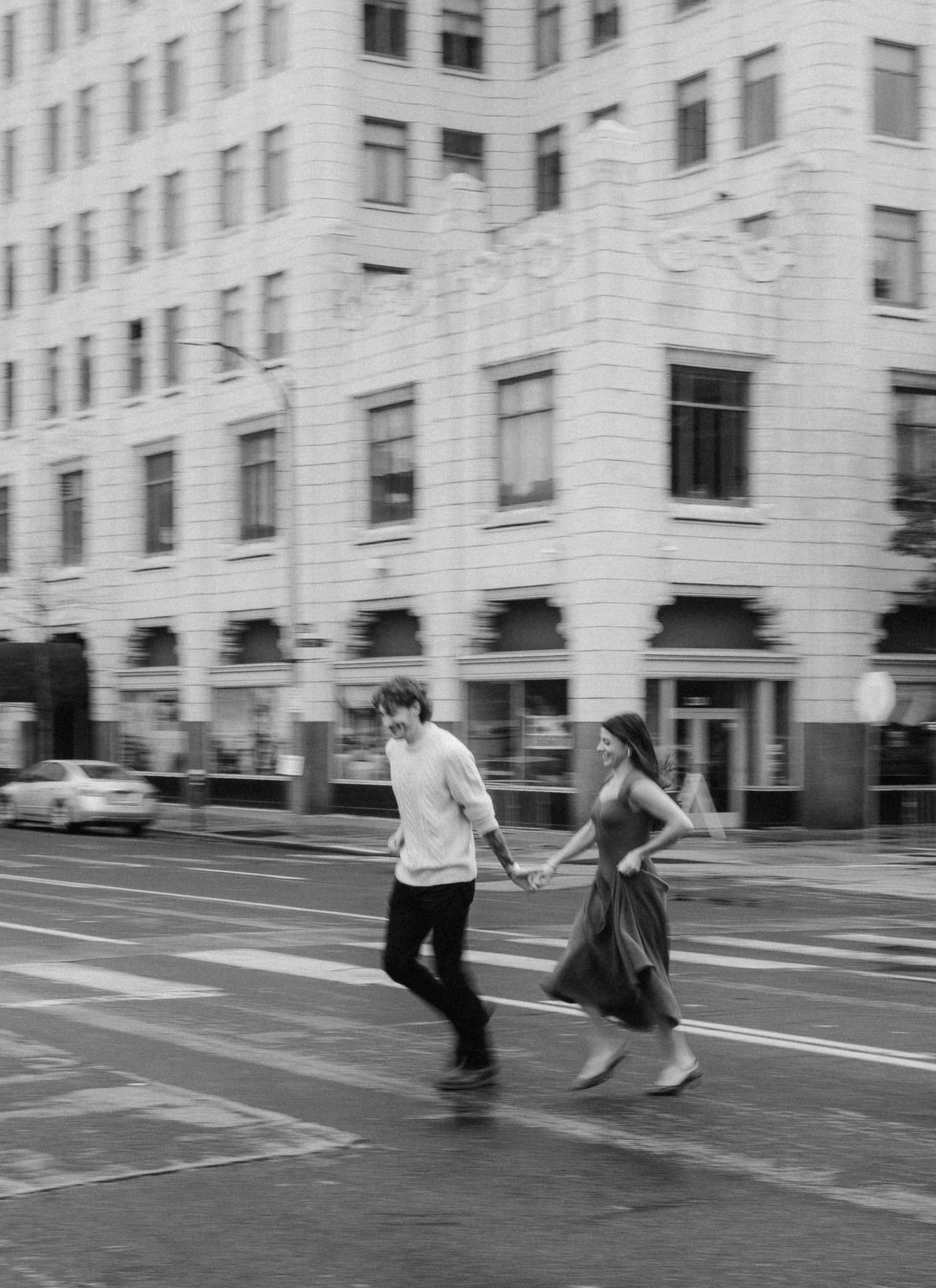 A couple runs across a downtown street in an urban engagement photoshoot.