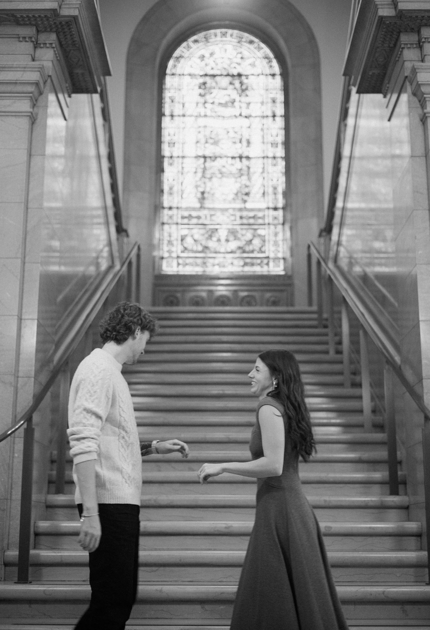 A couple laughs in front of a grand staircase during an engagement photoshoot at a library.