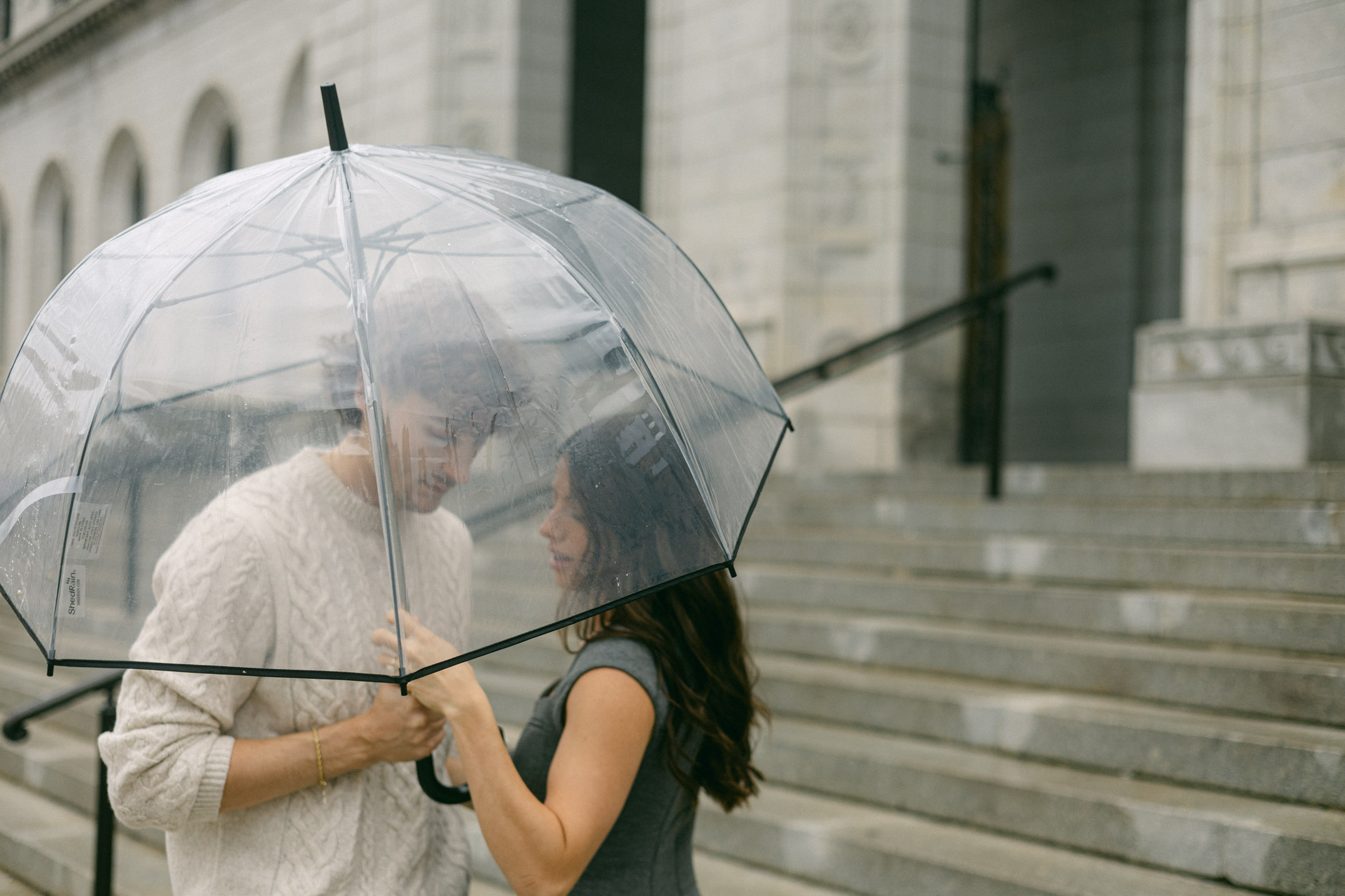 A couple under a clear umbrella on the steps of a downtown library.