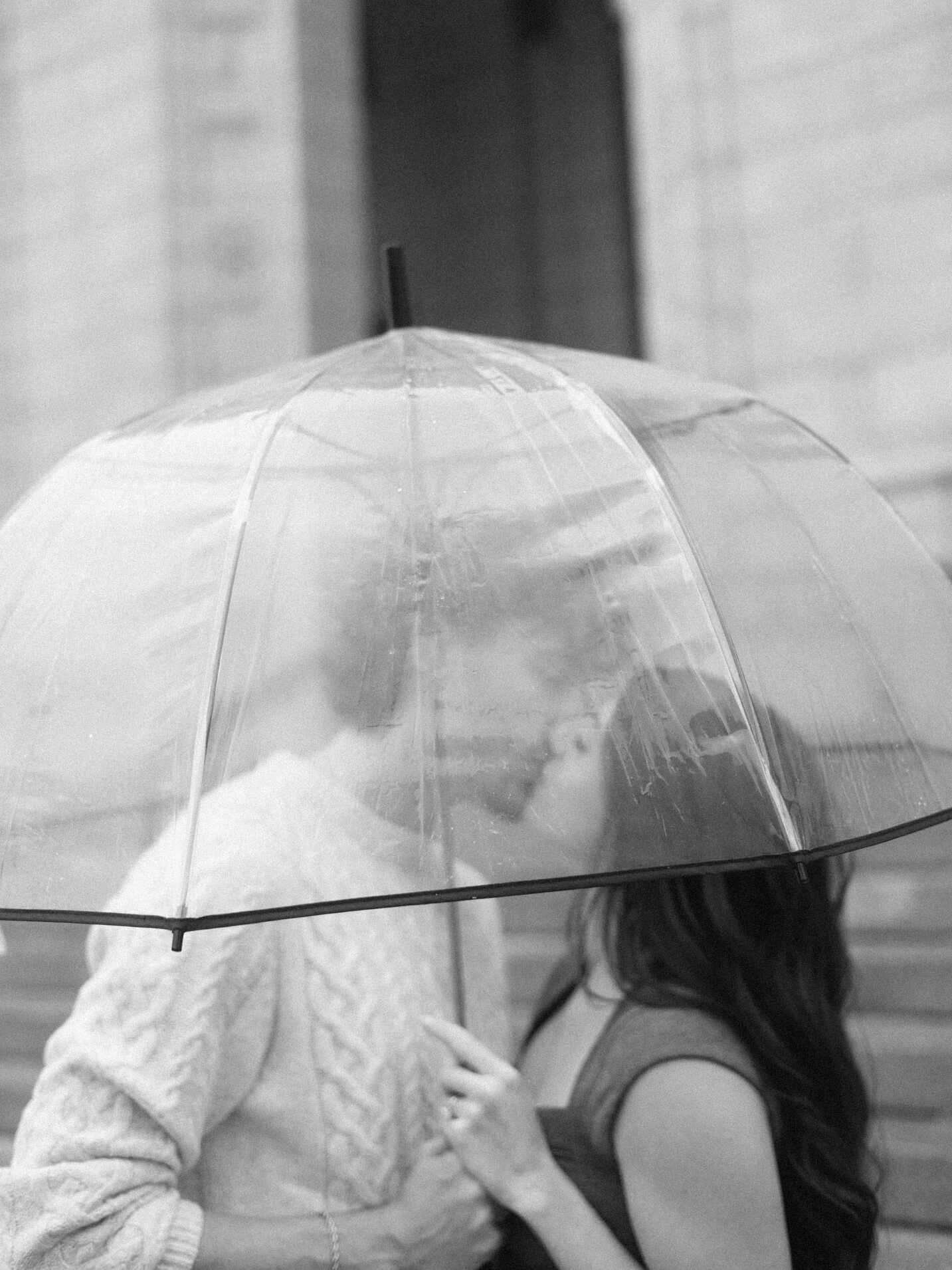 A couple kiss under a clear umbrella in a filmy engagement shoot.