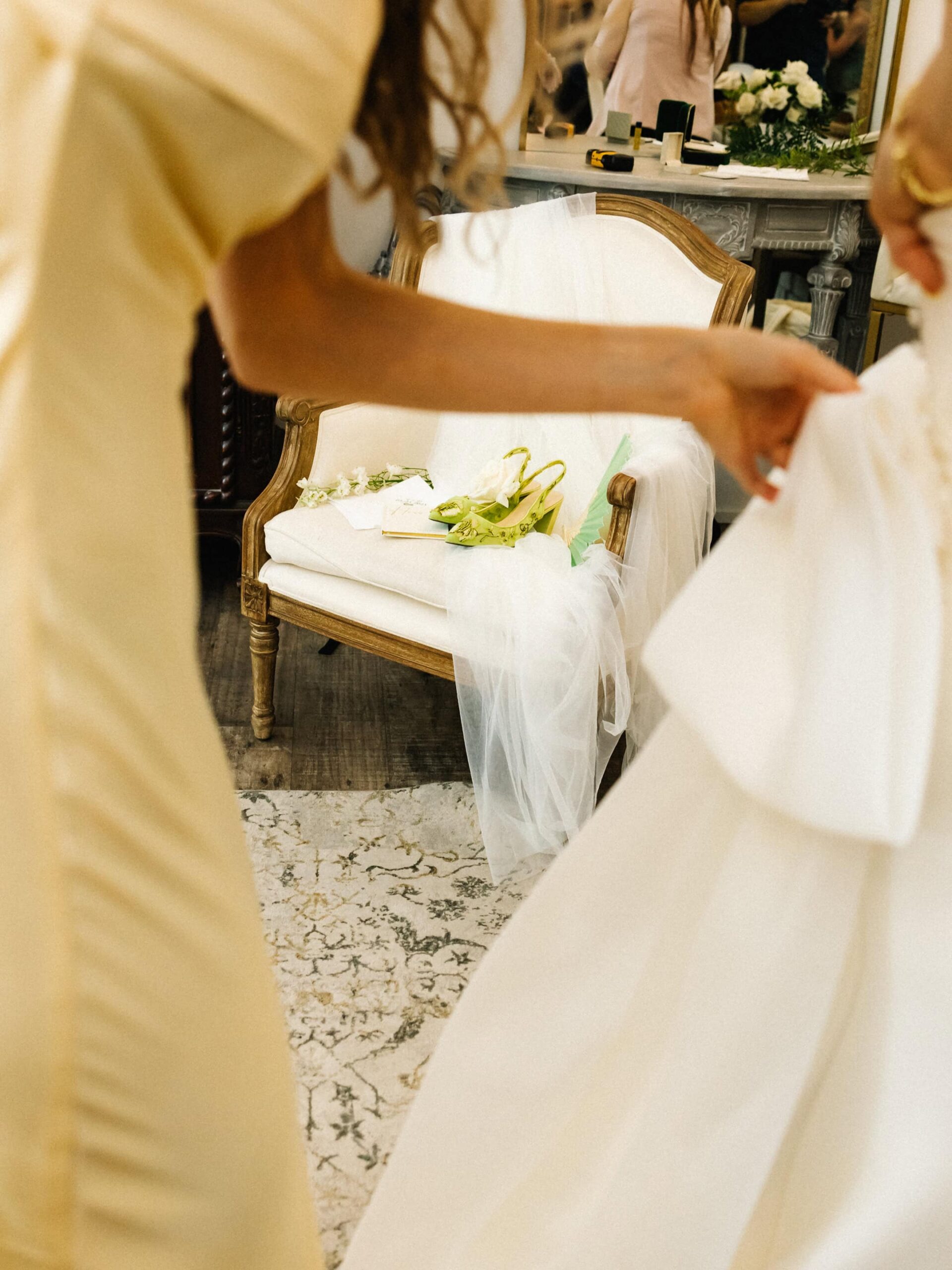 A documentary-style photo of bridal accessories on chair shot through the foreground of a bridesmaid helping the bride adjust her dress.