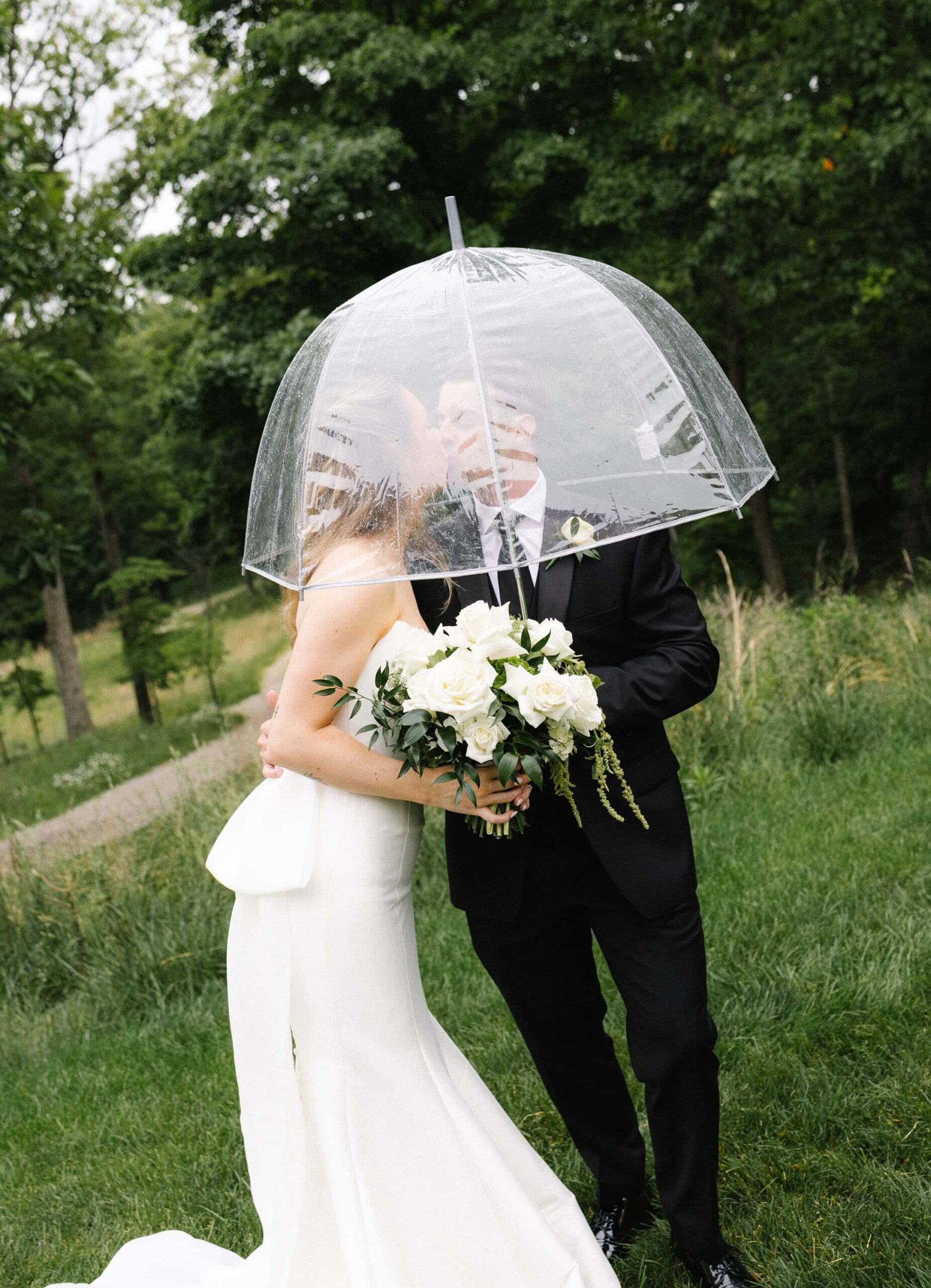 A bride and groom kiss outdoors under a clear umbrella in a documentary photography style.