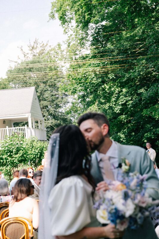 A bride and groom kiss outdoors at a wedding ceremony, with guests seated in the background and greenery surrounding the area.