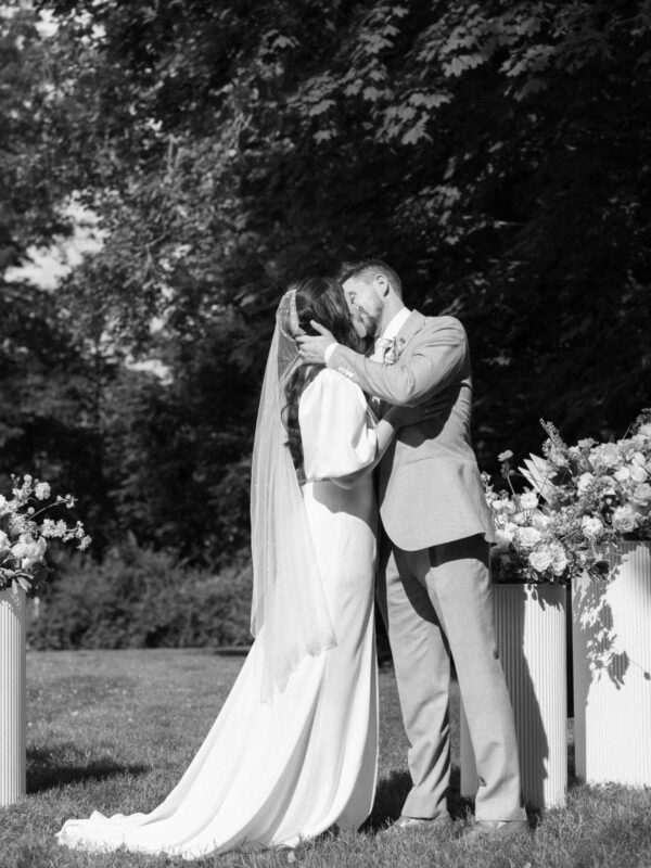 A bride and groom share a kiss outdoors during their wedding ceremony, surrounded by floral arrangements.