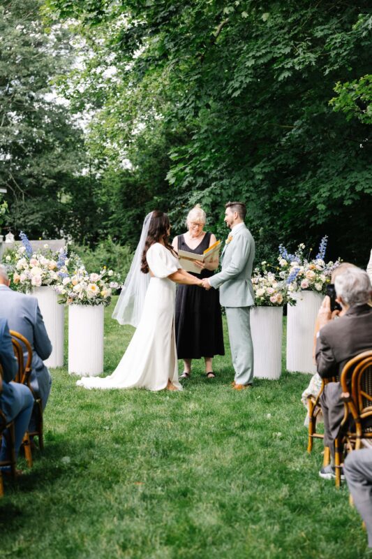 A couple stands facing each other, holding hands, during an outdoor wedding ceremony with an officiant and floral arrangements, surrounded by seated guests.