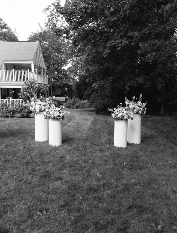Four white columns with flower arrangements stand on a grassy lawn, with a house and trees in the background. The image is in black and white.