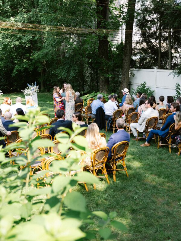 Wedding guests sit on wooden chairs arranged outdoors for an event, with greenery and trees in the background.