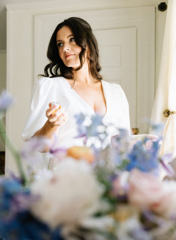 A bride stands indoors near a window, holding perfume bottle, with blurred flowers in the foreground.
