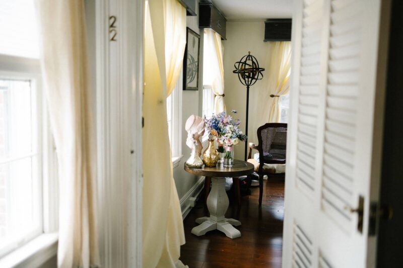 A sunlit room with sheer curtains, a round table holding vases and bouquets, wooden floors, and a chair near the window.