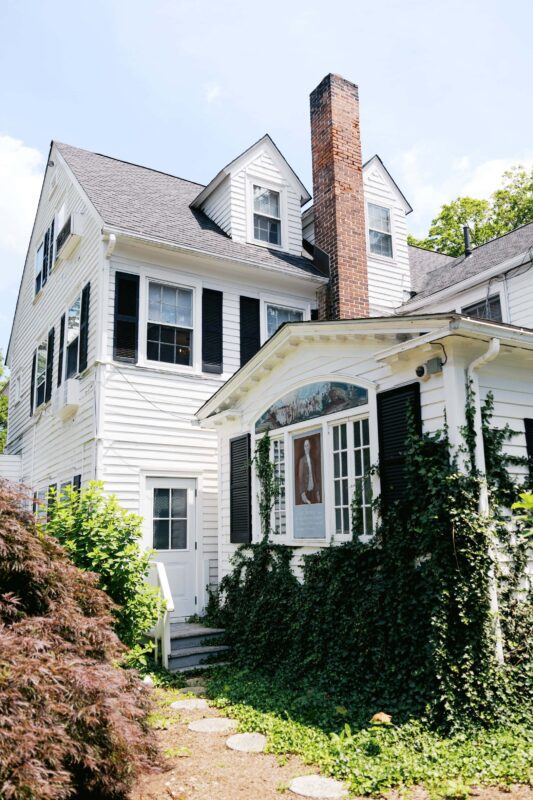 A white two-story house with black shutters, a brick chimney, and ivy growing up the front porch, surrounded by greenery on a sunny day.