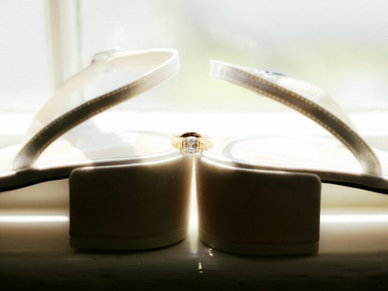 An egagement ring is placed between the heels of two white slingback wedding shoes, which are positioned back to back on a windowsill in natural light.