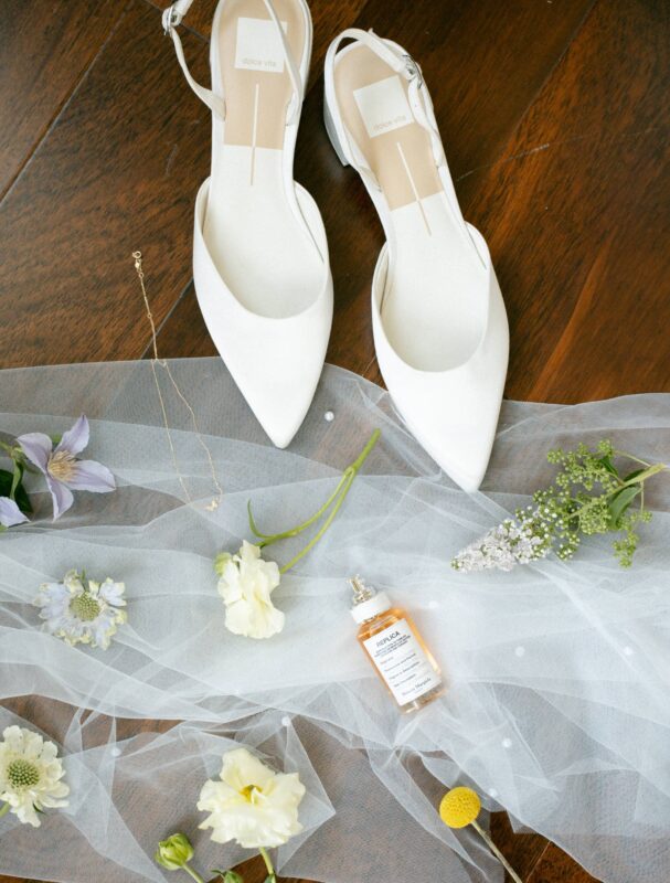 Flat lay of white pointed-toe bridal shoes, a sheer veil with small flowers, a delicate necklace, and a perfume bottle arranged on a wooden floor.
