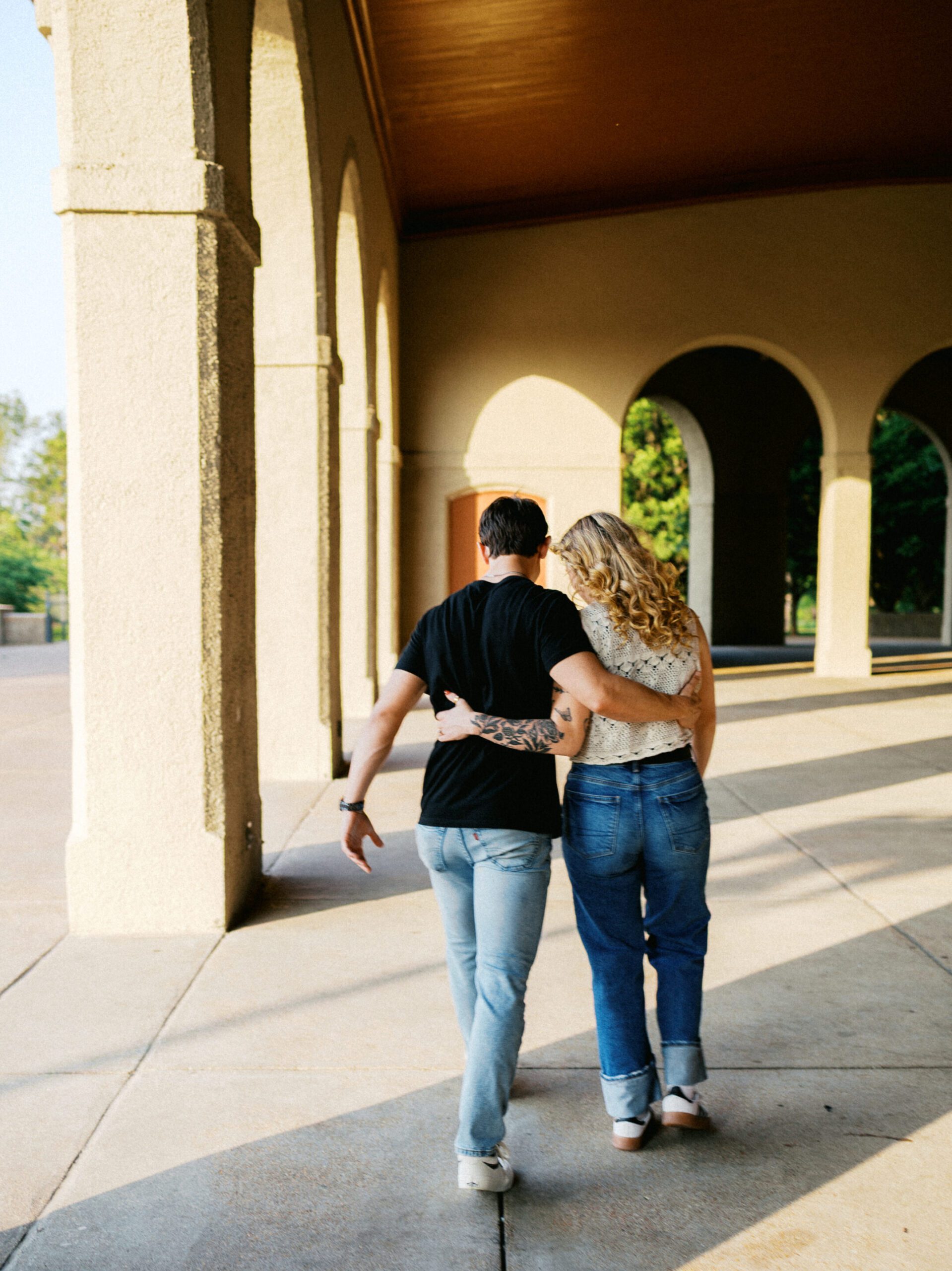A couple in jeans walk with arms around waists in an arched structure with shadowed light.