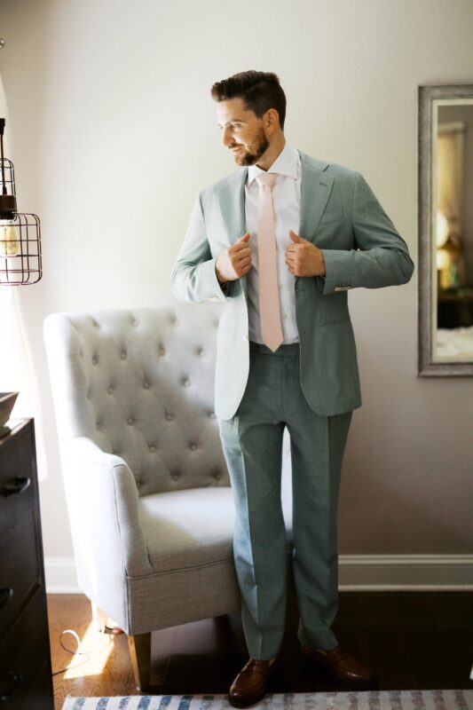 A groom in a light blue suit with a pale pink tie stands next to a gray armchair, adjusting his jacket and looking to the side.