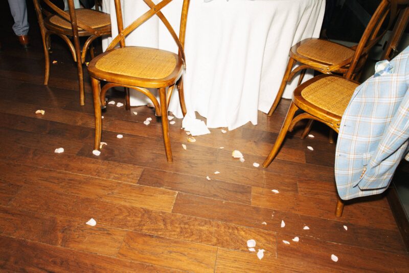 Scattered white flower petals are on a wooden floor around a dining table with wooden chairs; a plaid jacket hangs on one chair.