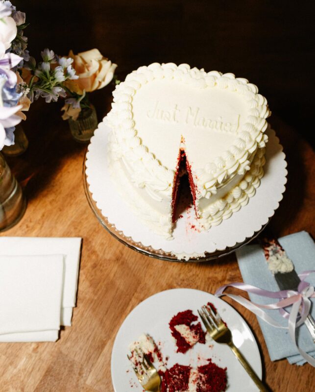 Heart-shaped wedding cake with “Just Married!” written on top, a slice cut out and served on a plate with two forks, flowers and napkins nearby on a wooden table.