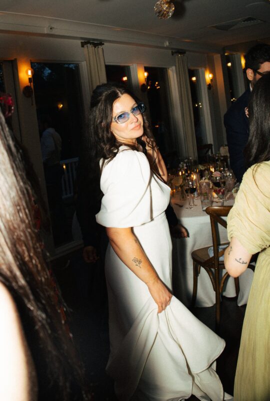 A bride in a white dress and blue sunglasses looks over her shoulder and smiles at a reception with tables, lights, and guests in the background.