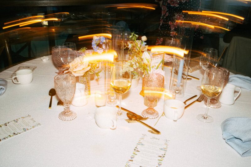 A round table set with glasses, cups, cutlery, menus, and floral centerpieces, with blurred yellow lights creating streaks across the image.