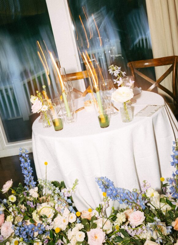 A round table covered with a white cloth, decorated with flowers and lit candles with light trails, sits in front of a window and two wooden chairs.