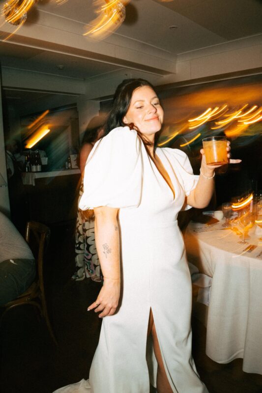 A bride holds a drink and smiles while dancing at an indoor reception, with warm, blurred lights in the background.