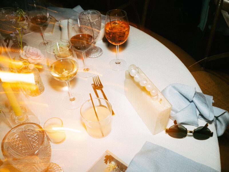A round table set with drinks, glassware, a fork and knife, a white clutch with pearls, sunglasses, and napkins under soft, warm lighting.