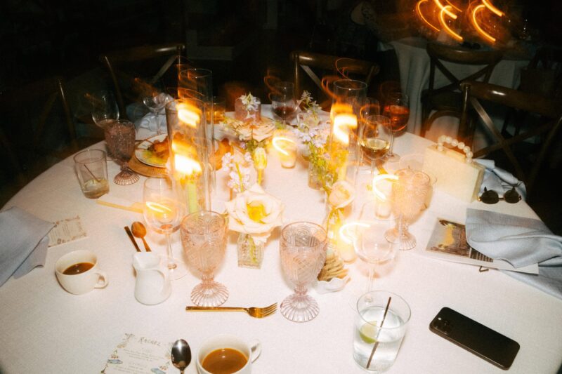 A round dinner table set with drinks, candles, glasses, flowers, and plates; some candles are blurred, indicating motion or a long exposure.