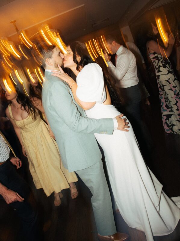 A couple in formal attire kisses on a dance floor surrounded by other people, with motion blur and warm lights creating a dynamic effect.