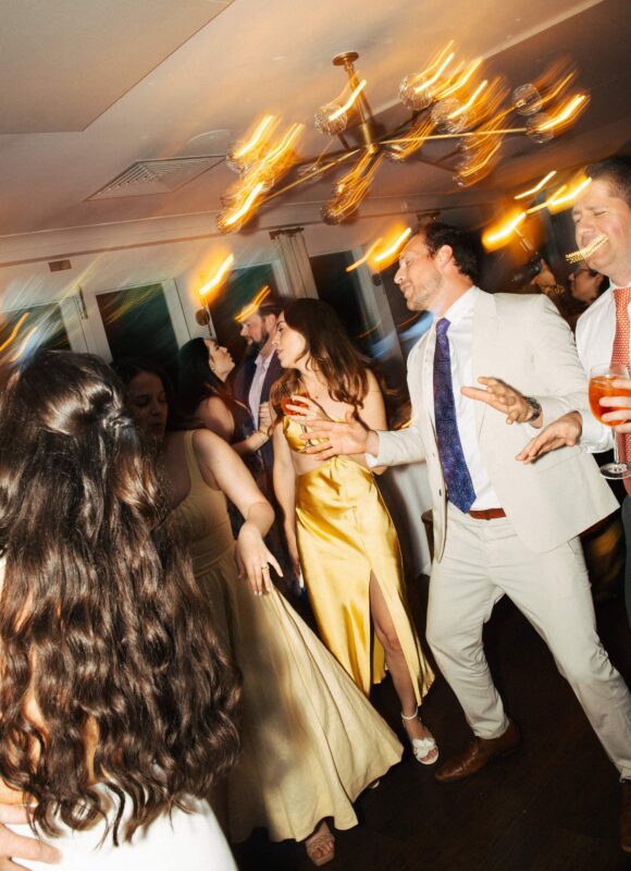 People dressed in formal attire dance and socialize at an indoor reception with warm lighting and blurred motion from moving chandelier lights.