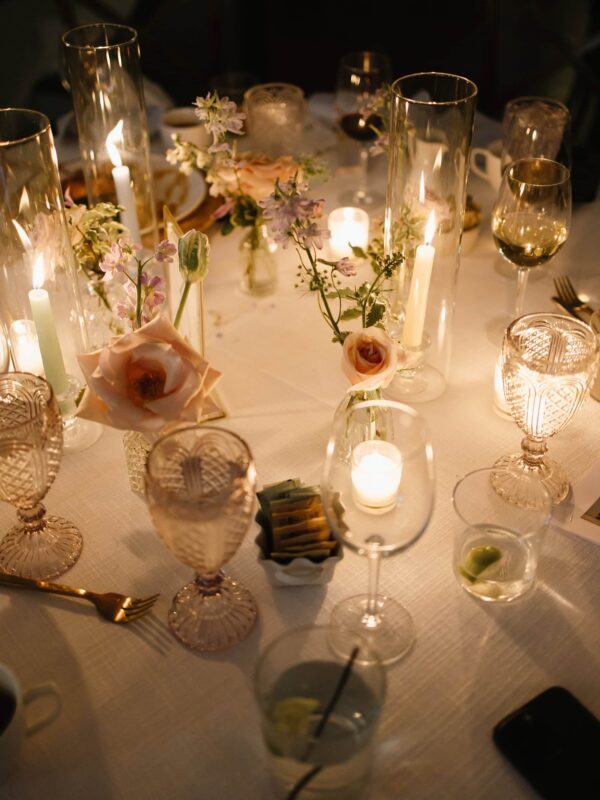 A round table set with glassware, candles, and small floral arrangements, creating a softly lit and elegant dinner setting.