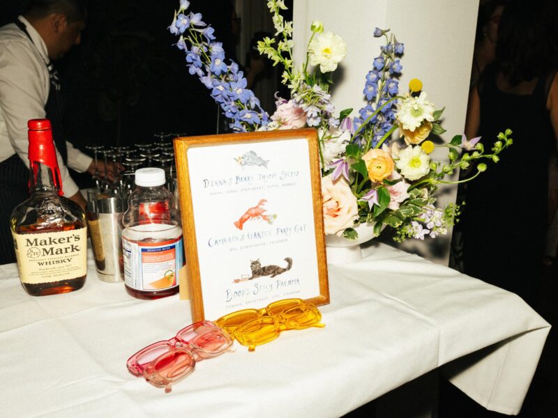 A table displays a bottle of Maker’s Mark, grenadine, pink and yellow sunglasses, a framed cocktail menu, and a floral arrangement.
