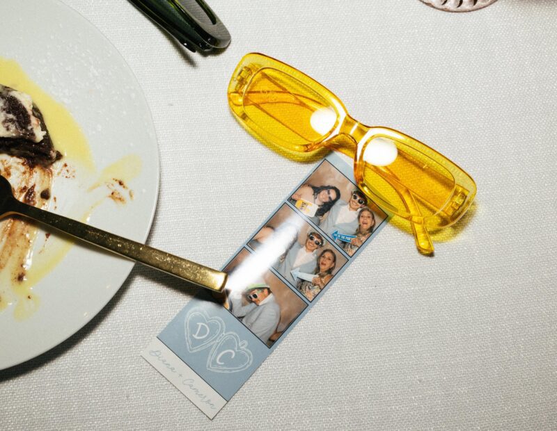 A pair of yellow sunglasses and a photo booth strip are on a white tablecloth next to a dessert plate with a gold spoon.
