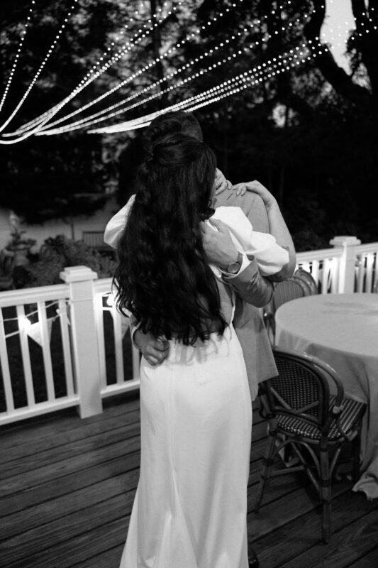 A couple embraces on a wooden deck at night under string lights, with empty chairs and a table nearby.