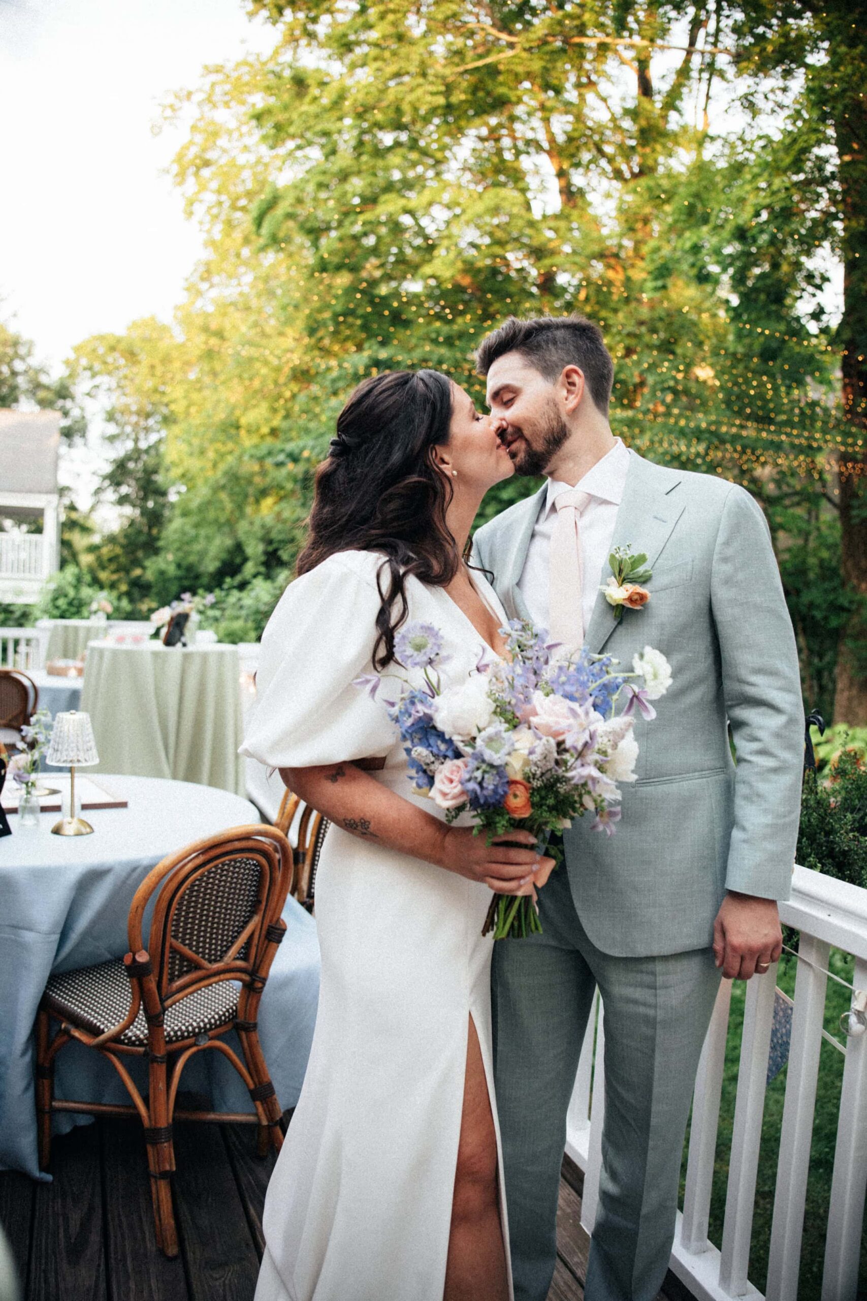 A bride and groom kiss on a porch with trees and golden light in the background.