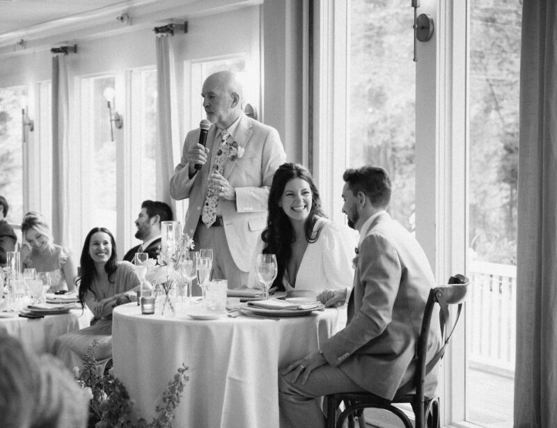An older man stands with a microphone giving a speech to a seated bride and groom at their wedding reception, while guests listen and smile.