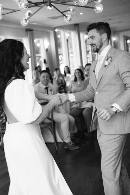 A bride and groom dance together at their wedding reception while seated guests watch and smile in the background.