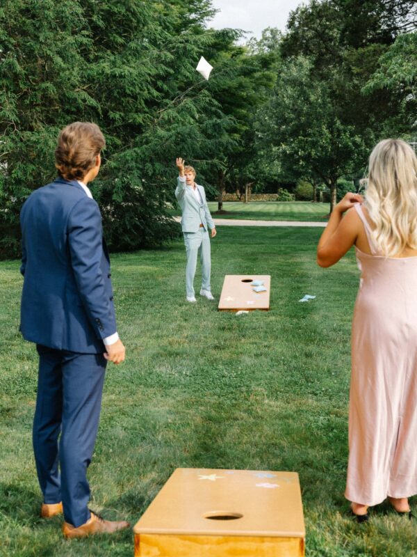 Three people dressed formally play cornhole on a grassy lawn, with one person tossing a bean bag toward the board.