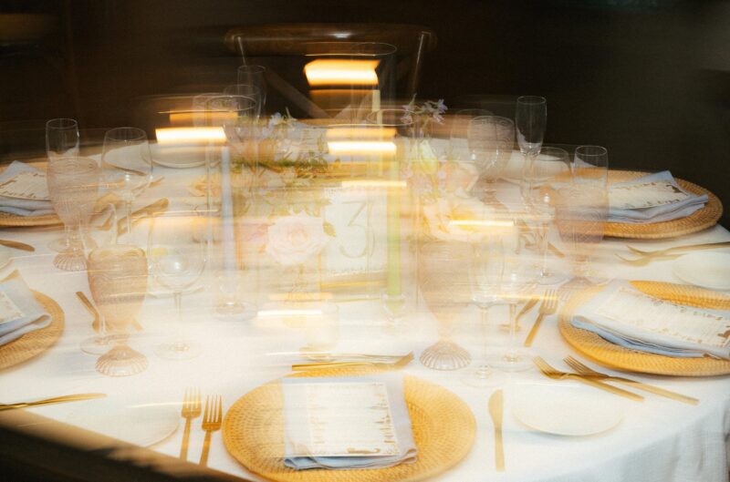 A blurred image of a round dining table set with plates, glasses, cutlery, woven chargers, and a floral centerpiece with a table number.