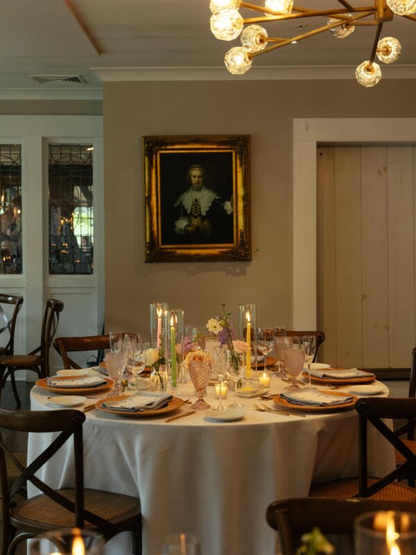 Round dining table set with plates, glasses, and candles beneath a modern chandelier, with a framed portrait on the beige wall in the background.