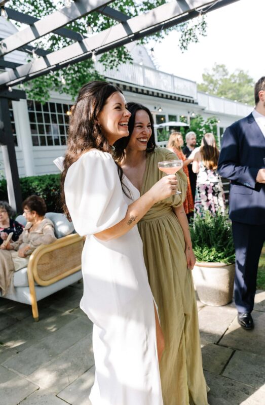 A bride and her friend smile and hold drinks at an outdoor cocktail hour, with other guests socializing in the background.