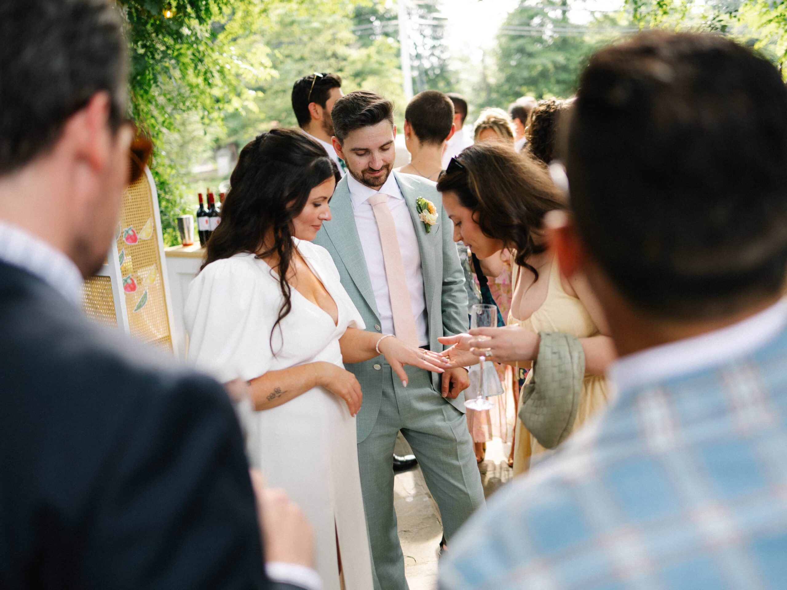 A bride shows her ring to guests at an outdoor wedding reception, with the groom standing beside her and people gathered around.