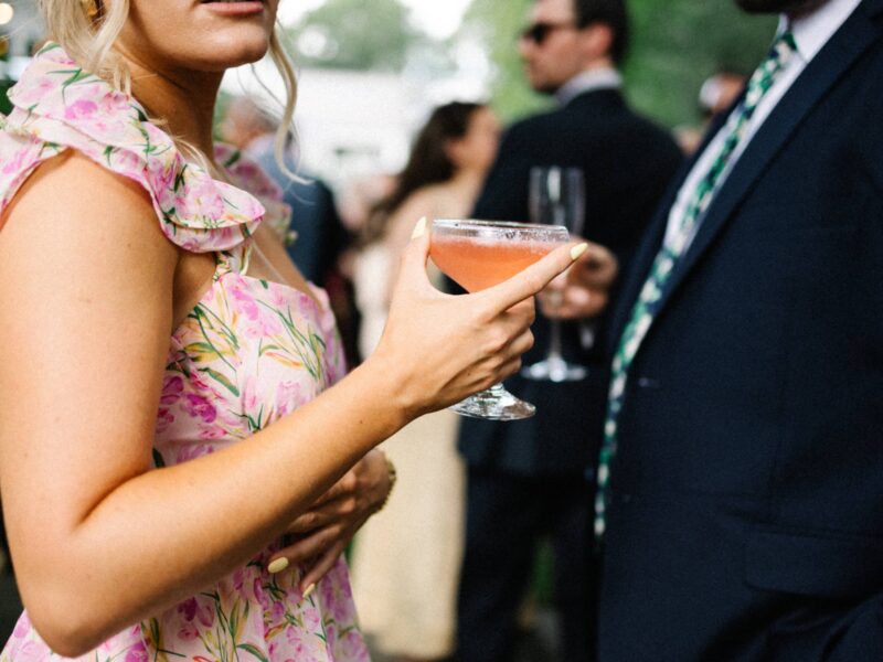 A maid of honor in a floral dress holds a pink cocktail while standing among other wedding guests at an outdoor cocktail hour.