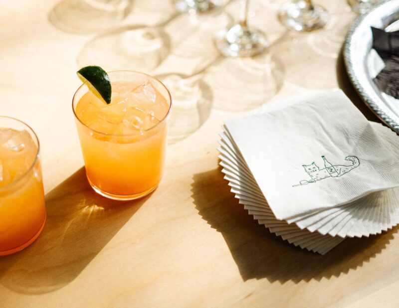 Two orange cocktails with ice and lime wedges sit on a table next to a stack of white napkins with a cat illustration.